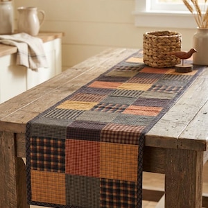 May include: A rectangular table runner with a patchwork design. The runner features squares in shades of orange, brown, yellow, and black. The runner is placed on a rustic wooden table. A woven basket and decorative bird are on the table.