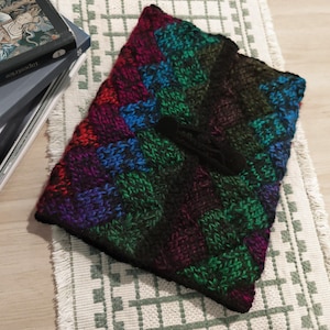 May include: A rectangular, hand-knitted book cover featuring a diagonal, multi-colored pattern in shades of deep purple, vibrant blue, emerald green, and black. The cover includes a black knitted strap closure centered on the front panel.