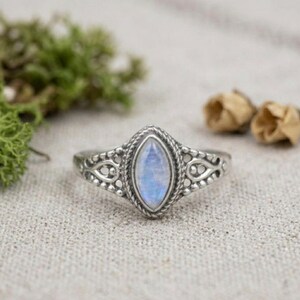 Könnte beinhalten: Ein silberner Ring mit einem hellblauen, marquiseförmigen Edelstein. Der Ring hat ein aufwendiges, detailliertes Design um den Stein und entlang des Bandes. Der Ring ist vor einem neutralen Hintergrund mit Grün und getrockneten Blumen platziert.