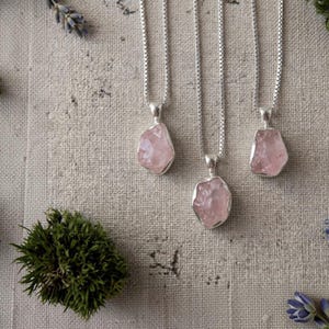 Op de afbeelding: Drie zilveren kettingen met ruwe, lichtroze rozenkwarts hangers. Elke hanger hangt aan een fijne zilveren ketting en toont de natuurlijke, onregelmatige vorm en de doorschijnende textuur van het ongeslepen edelsteenmateriaal.