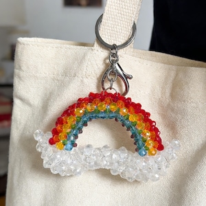 Könnte beinhalten: Ein bunter Schlüsselanhänger in Regenbogenform aus Perlen in Rot, Orange, Gelb, Grün und Blau. Der Regenbogen sitzt auf einer Wolke aus klaren Perlen. Der Schlüsselanhänger ist an einem silbernen Verschluss und einer cremefarbenen Tragetasche befestigt.