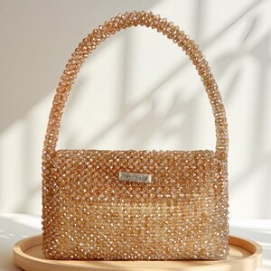 Könnte beinhalten: Handgefertigte, rechteckige Handtasche, bedeckt mit kleinen, funkelnden, bernsteinfarbenen Perlen. Die Tasche hat einen passenden Perlenhenkel und ein kleines silbernes Etikett mit der Aufschrift "handmade". Die Tasche steht auf einem runden, hellen Holztablett.