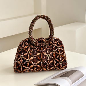 Puede incluir: Un bolso de mano marrón con cuentas y un asa redondeada. El bolso presenta un patrón floral hecho de cuentas marrones y doradas. El asa también está cubierta de cuentas. El bolso está sobre una superficie blanca.