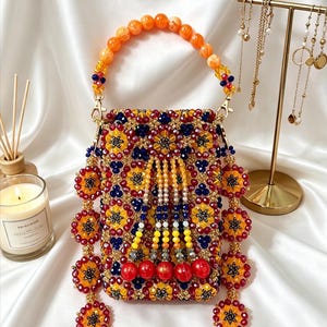 Könnte beinhalten: Eine mit Perlen besetzte Handtasche mit einem lebendigen Design. Die Tasche hat einen Griff aus orangefarbenen Perlen und ist mit gelben, roten und blauen Perlen verziert. Dekorative Perlenquasten hängen von unten. Ein goldener Schmuckständer und eine Kerze sind im Hintergrund.
