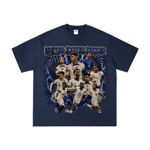 May include: A navy blue short-sleeve t-shirt featuring a graphic print of the United States soccer team. The design includes multiple players in white jerseys, blue lightning bolts, and wings, with the text UNITED STATES SOCCER at the top.