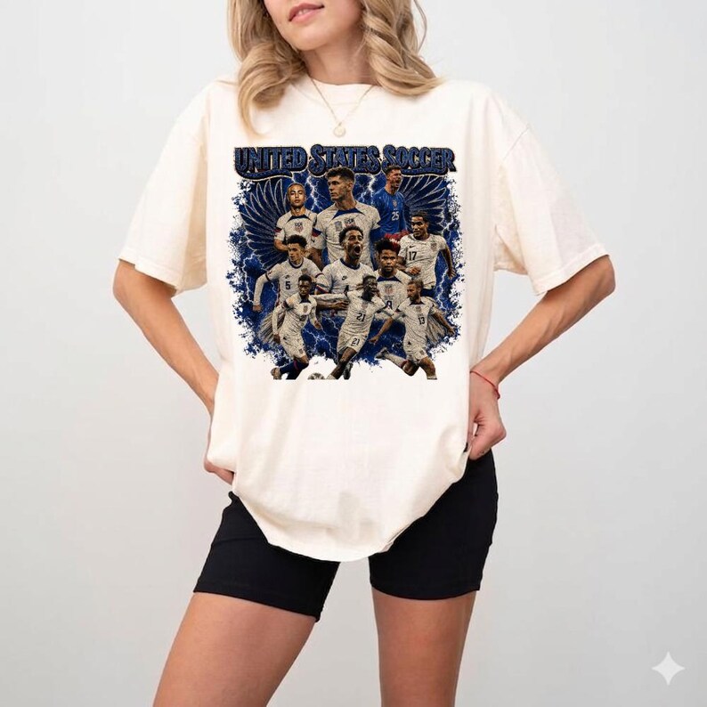 May include: An off-white short-sleeve t-shirt featuring a graphic print of the United States Soccer team. The design includes multiple player portraits, blue wing accents, and the text UNITED STATES SOCCER in a bold, dark blue font.