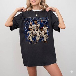 May include: A black short-sleeve t-shirt featuring a graphic print of the United States soccer team. The design includes multiple players in white jerseys, blue lightning bolts, and the text United States Soccer in a bold blue font at the top.