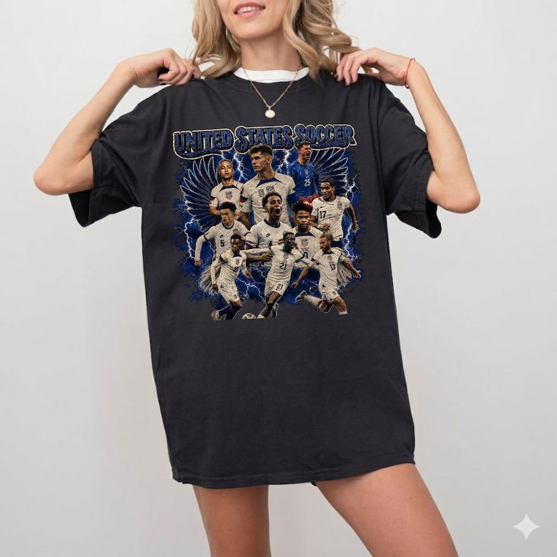 May include: A black short-sleeve t-shirt featuring a graphic print of the United States soccer team. The design includes multiple players in white jerseys, blue lightning bolts, and the text United States Soccer in a bold blue font at the top.