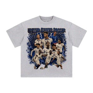 May include: A heather gray short-sleeve t-shirt featuring a graphic print of the United States Soccer team. The design includes multiple players in white jerseys, blue lightning bolts, and wings, with the text UNITED STATES SOCCER at the top.