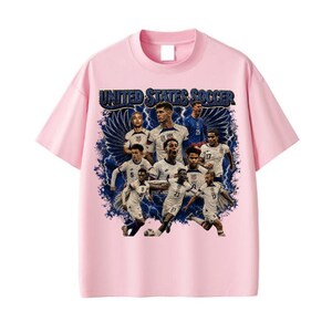 May include: A light pink short-sleeve t-shirt featuring a graphic print of the United States Soccer team. The design includes multiple players in white jerseys, dark blue lightning bolts, and wings, with the text UNITED STATES SOCCER at the top.