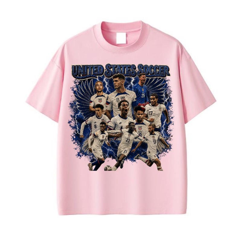 May include: A light pink short-sleeve t-shirt featuring a graphic print of the United States Soccer team. The design includes multiple players in white jerseys, dark blue lightning bolts, and wings, with the text UNITED STATES SOCCER at the top.