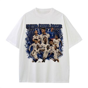May include: A white short-sleeve t-shirt featuring a graphic print of the United States soccer team. The design includes multiple players in white jerseys, blue lightning effects, and the text UNITED STATES SOCCER in a bold blue font at the top.