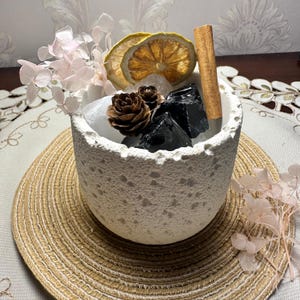 May include: A white textured cylindrical container filled with decorative elements including two dried lemon slices, a cinnamon stick, a small brown pinecone, clear quartz-like stones, black obsidian-like stones, and pale pink dried hydrangea flowers.