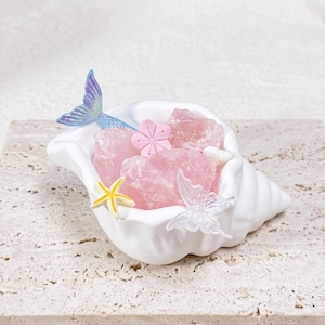 May include: A white ceramic shell-shaped dish filled with raw pink rose quartz crystals. The dish is decorated with a small iridescent blue mermaid tail, a pink flower, a yellow starfish, and a translucent butterfly figurine.
