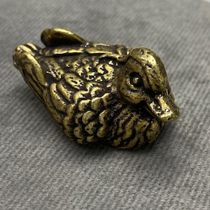 May include: A small, detailed brass duck figurine. The duck is in a resting position, with textured feathers and a detailed beak. The brass has a slightly aged, antique appearance, with variations in color.