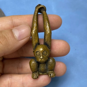 May include: A bronze monkey figurine with long arms looped at the top. The monkey has a detailed face and is in a hanging position. The figurine is likely a decorative item or collectible, possibly made of brass or a similar metal.