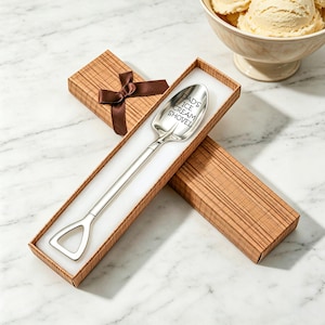 Puede incluir: Una pala de helado plateada con el texto "DAD'S ICE CREAM SHOVEL" grabado en la cuchara, presentada en una caja de regalo marrón con una cinta marrón. La pala tiene un mango largo y una cuchara en forma de pala.