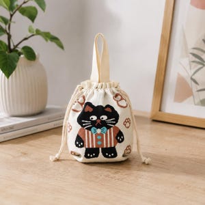 May include: A cream-colored knit drawstring pouch featuring a central graphic of a black cat wearing a brown striped shirt and a light blue bow tie. The bag has a single top handle and is decorated with small brown paw print patterns throughout.