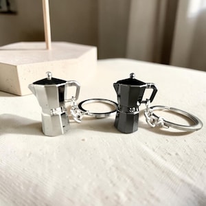 May include: Two metal keychains shaped like moka pot coffee makers. One keychain is finished in a polished silver tone, while the other features a dark, gunmetal grey finish. Each miniature coffee pot is attached to a standard circular metal ring.