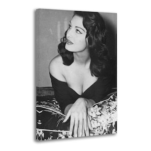 May include: Black and white portrait of a woman with dark, wavy hair, wearing a low-cut black top. She is reclining on a floral patterned pillow, looking off to the side. The image is a vintage style photograph.