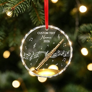 Peut inclure: Une décoration circulaire en acrylique transparent ornée d'un motif de flûte dorée, de notes de musique blanches et de portées. Le texte indique CUSTOM TEXT, Name et 2026. Elle est suspendue par un ruban rouge fin sur un fond de sapin flou.