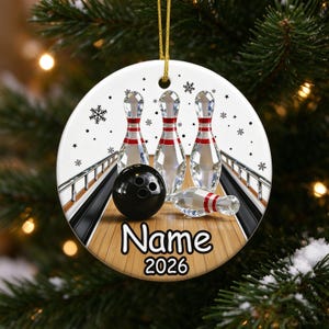 May include: A round white ceramic ornament featuring a bowling alley graphic with three upright crystal bowling pins, one fallen pin, and a black bowling ball. The text Name and 2026 are printed in black below the pins, with snowflakes.