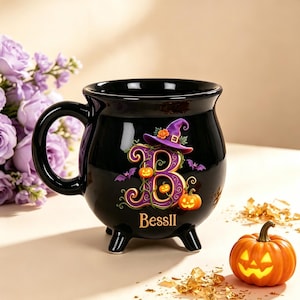 Pode incluir: Uma caneca preta em forma de caldeirão com três pés, apresentando uma letra B grande roxa decorada com vinhas, pequenas abóboras laranja e um chapéu de bruxa roxo. O nome BESSII está impresso em dourado abaixo da letra com morcegos roxos.