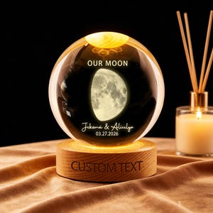 Op de afbeelding: Een heldere kristallen bol met een maanbeeld erin, rustend op een houten voet. De bol is van binnenuit verlicht en toont de tekst "OUR MOON", namen en een datum. De houten voet heeft de tekst "CUSTOM TEXT" gegraveerd.