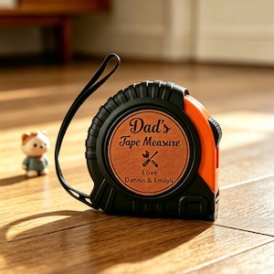 May include: A black and orange tape measure with the text "Dad's Tape Measure" and "Love, Dannis & Emilyli" on a brown circular label. The tape measure has a black strap and a small figurine is in the background.