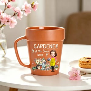 Può includere: Una tazza in ceramica color terracotta a forma di vaso da fiori, con il testo GARDENER of the Year e NAME. Il design presenta un personaggio dei cartoni animati con un grembiule giallo, circondato da fiori in vaso, attrezzi e colibrì.