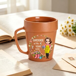 Può includere: Una tazza in ceramica color terracotta a forma di vaso da fiori, con un'illustrazione stampata di un giardiniere, una ghirlanda floreale e il testo "GARDENER of the YEAR NAME". La tazza presenta un bordo largo, un manico robusto e una finitura opaca.