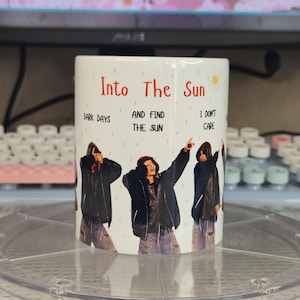 Pode incluir: Caneca de cerâmica branca com o texto "Into The Sun" e as frases "Dark Days", "And Find The Sun" e "I Don't Care". A caneca apresenta um design de três pessoas em casacos pretos e jeans.