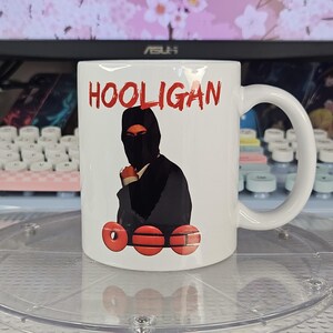 May include: White ceramic mug featuring the word "HOOLIGAN" in red, above an illustration of a person in a black balaclava. Below the illustration are three red circles with horizontal lines.
