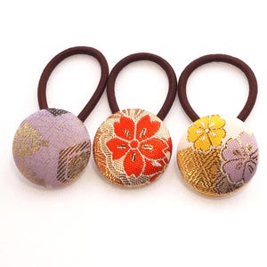 May include: Three decorative hair ties with brown elastic bands. Each tie features a round, fabric-covered button with a floral design. The buttons are in shades of lavender, orange, and yellow, with gold accents. The background is white.