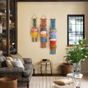 Op de afbeelding: Drie verticale macramé wandhangers met geïntegreerde vakken voor het opbergen van baseballpetten. De linkerhanger is saliegroen, de middelste roestoranje en de rechter is blauwgroen, elk met meerdere kleurrijke baseballpetten erin.