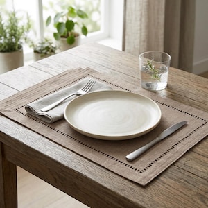 May include: A dining table setting with a beige placemat, a white plate, a folded gray napkin, a fork, and a knife. A glass of water with a sprig of rosemary is also present. The table is made of wood.