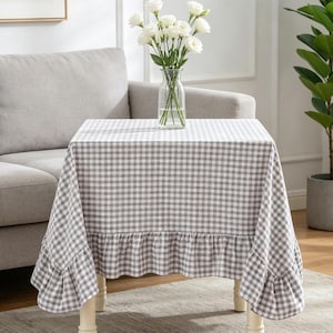 May include: A small, square table covered with a gray and white checkered tablecloth. The tablecloth has a ruffled edge. A clear glass vase with white flowers sits on the table. The table has white legs.