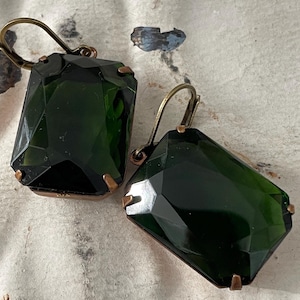 May include: A pair of dark green glass earrings with a faceted design. The earrings have a bronze-colored metal hook.
