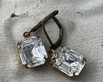 May include: A pair of gold tone earrings with clear, faceted, square-shaped crystals. The earrings have a lever back closure.