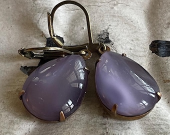 May include: A pair of vintage earrings featuring teardrop-shaped, lavender-colored stones. The stones are set in a gold-toned metal frame with a lever-back closure. The earrings have a classic, elegant design.