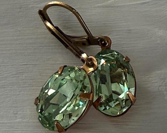 May include: A pair of leverback earrings featuring oval-shaped, faceted light green crystals set in an antique bronze-toned metal frame. The crystals have a brilliant cut with multiple reflective surfaces, held securely by four small metal prongs.