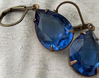 May include: A pair of blue teardrop-shaped earrings with antique brass settings. The earrings have a faceted design and are suspended from a hook.