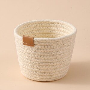 May include: A small, round, cream-coloured woven basket with a brown leather accent. The basket is made of thick, braided rope and has a cylindrical shape. The leather accent is a small rectangular patch attached to the side.