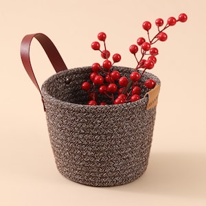Puede incluir: Una pequeña cesta marrón tejida con un asa de cuero burdeos y una ramita de bayas rojas. La cesta tiene un aspecto texturizado y una pequeña etiqueta de cuero. Ideal para la decoración del hogar o el almacenamiento.