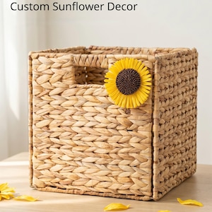 May include: Woven storage basket with custom sunflower decor. The square basket is made of a light brown woven material. A yellow sunflower is attached to the front. The text "Woven Storage Basket" and "Custom Sunflower Decor" are at the top. The text "Eco-friendly Material" is at the bottom.