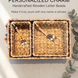 May include: Three rectangular woven seagrass baskets nested together. Two baskets contain personalized charms made of wooden letter beads in various colors, including yellow, orange, black, and white, with white cord accents. Text reads: PERSONALIZED CHARM Handcrafted Wooden Letter Beads Make it truly yours with any name or phrase.