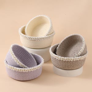 Puede incluir: Tres cestas decorativas tejidas en crema, lavanda y topo. Cada cesta está adornada con una fila de pequeñas perlas blancas alrededor del borde. Las cestas están anidadas y dispuestas sobre pedestales blancos sobre un fondo neutro.