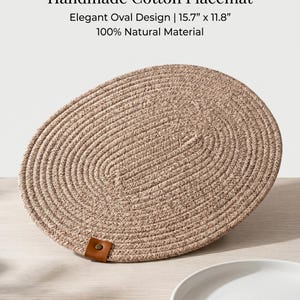 Può includere: Un sottopiatti ovale in cotone fatto a mano, di colore beige, con un design elegante. Il sottopiatti è realizzato in materiale naturale al 100% e presenta una trama intrecciata. Ha un'etichetta in pelle marrone attaccata. Il testo sull'immagine recita: "Handmade Cotton Placemat, Design ovale elegante | 40cm x 30cm, 100% Natural Material."