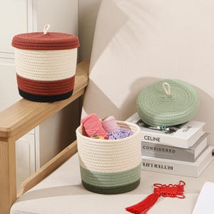 May include: A collection of woven rope baskets in various sizes and colors. One basket has a lid, another contains yarn, and a third is empty. The color scheme includes red, cream, green, and black.