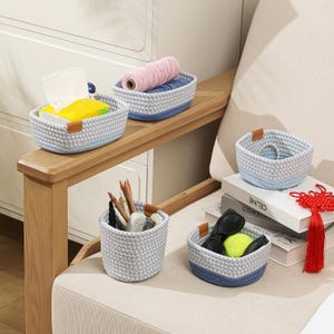 May include: A collection of small, woven rope baskets in various sizes and shapes. The baskets are primarily light gray with a blue base and a brown leather accent. Some baskets contain items like pencils, sunglasses, and yarn.